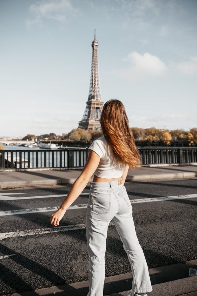 Woman in casual attire stands on a Paris bridge with the Eiffel Tower in the background.